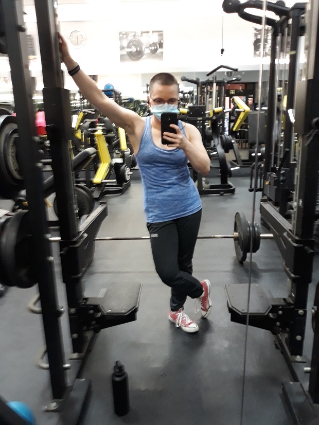 Mirror reflection of a woman standing in a squat rack in a gym. She has a very short buzzcut and has glasses and a surgical mask on. She is wearing a close fitting blue tank top, black training pants, and red Converse sneakers. In her left hand she is holding a phone with which she is taking the picture, and her right hand is raised and holding onto the squat rack. Behind her there is a loaded barbell on the floor.