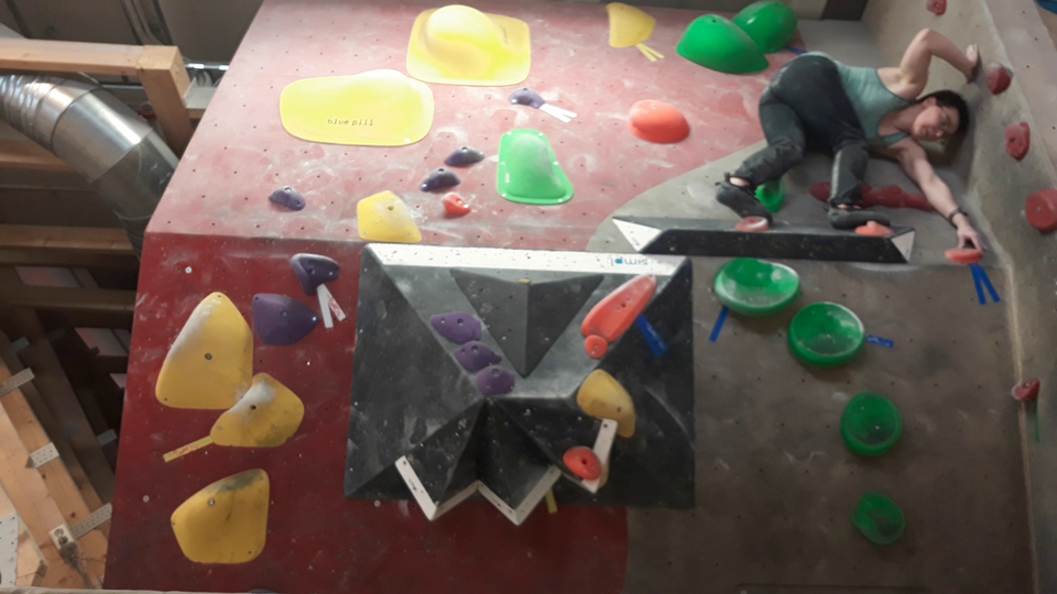 Two bouldering walls forming a corner. A woman is standing on two orange holds next to each other about 5-6 feet off the ground, with both her feet facing right. Her legs are slightly bent, and she is bending over to the right, with her body twisted so that she is touching another orange hold further on the right and a bit lower than her feet with her left hand, while her right arm is bent over her head as she is pushing against the perpendicular wall with her right hand.